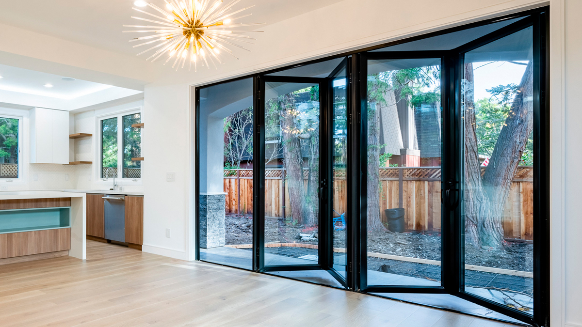 ประตูบานเฟี้ยมกระจกใสหลายบาน กรอบสีดำ กำลังเปิดออก ทำให้เห็นสวนหลังบ้านที่มีต้นไม้ใหญ่ รั้วไม้ และพื้นดิน ภายในห้องมีพื้นไม้สีอ่อน เคาน์เตอร์ครัวสีขาว และโคมไฟระย้าดีไซน์ทันสมัย, Multiple clear glass bifold doors with black frames are opening, revealing a backyard garden with large trees, a wooden fence, and soil. Inside the room, there is a light wood floor, a white kitchen counter, and a modern design chandelier.