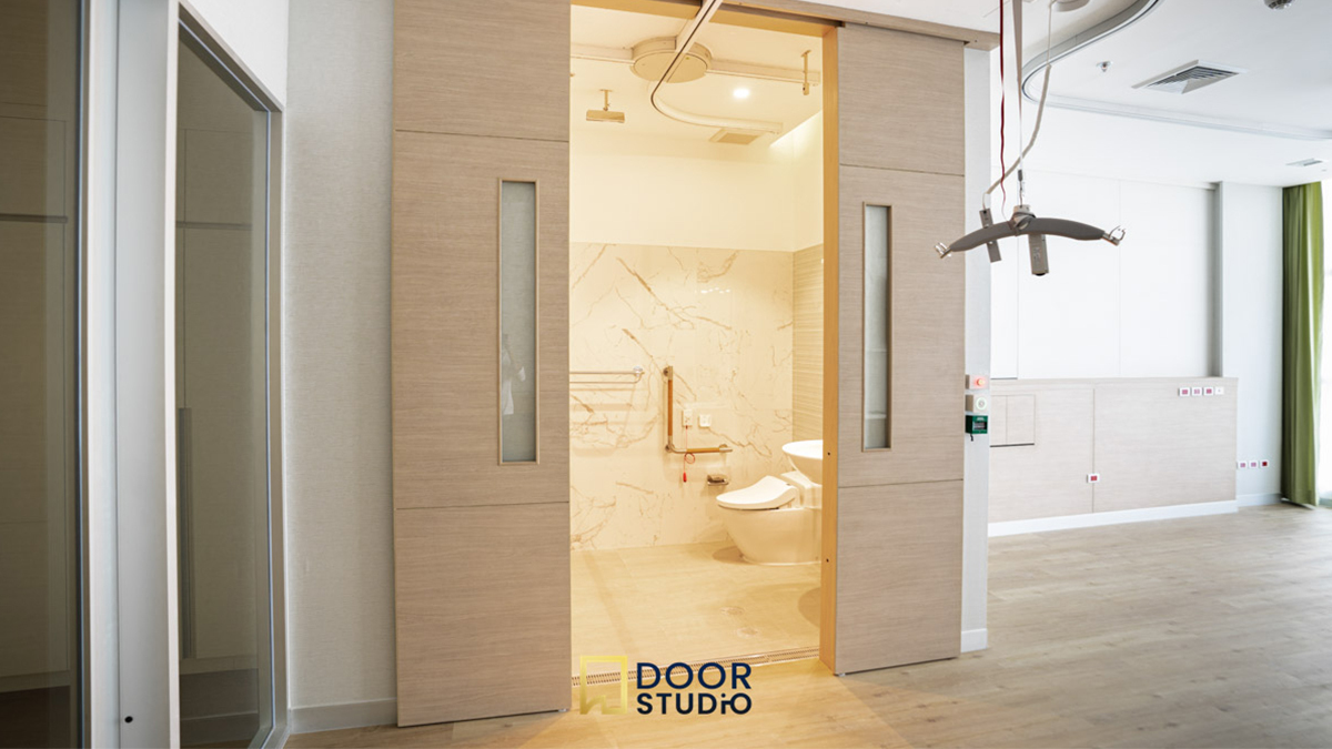 ประตูบานเลื่อนอัตโนมัติทำจากวัสดุสีอ่อน เปิดออกเล็กน้อย เผยให้เห็นภายในห้องน้ำที่มีสุขภัณฑ์สีขาว ผนังลายหินอ่อน และราวจับสำหรับผู้สูงอายุหรือผู้พิการ ติดตั้งในพื้นที่ที่ดูเหมือนโรงพยาบาลหรือสถานพยาบาล, Light-colored automatic sliding door slightly open, revealing a bathroom interior with white sanitary ware, marble-patterned walls, and grab bars for the elderly or disabled. Installed in what appears to be a hospital or healthcare facility.