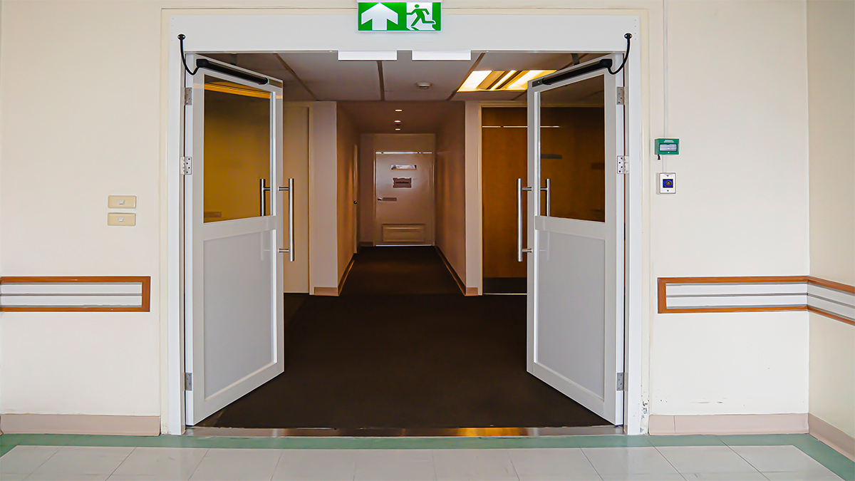 ประตูบานสวิงอัตโนมัติสองบานสีขาว เปิดค้างอยู่ เผยให้เห็นทางเดินภายในอาคารที่ดูเหมือนโรงพยาบาล มีปุ่มกดสีเขียวสำหรับเปิดประตูติดตั้งอยู่ข้างผนังด้านขวา เหนือประตูมีป้ายทางออกฉุกเฉินสีเขียวเรืองแสง พื้นเป็นกระเบื้องสีอ่อน และมีแถบกันกระแทกสีน้ำตาลคาดตามแนวกำแพง, Two white automatic swing doors are held open, revealing a hallway inside what appears to be a hospital. A green push button for opening the doors is mounted on the right wall. Above the doors is a glowing green emergency exit sign. The floor is light-colored tile, and brown bumper guards run along the walls.