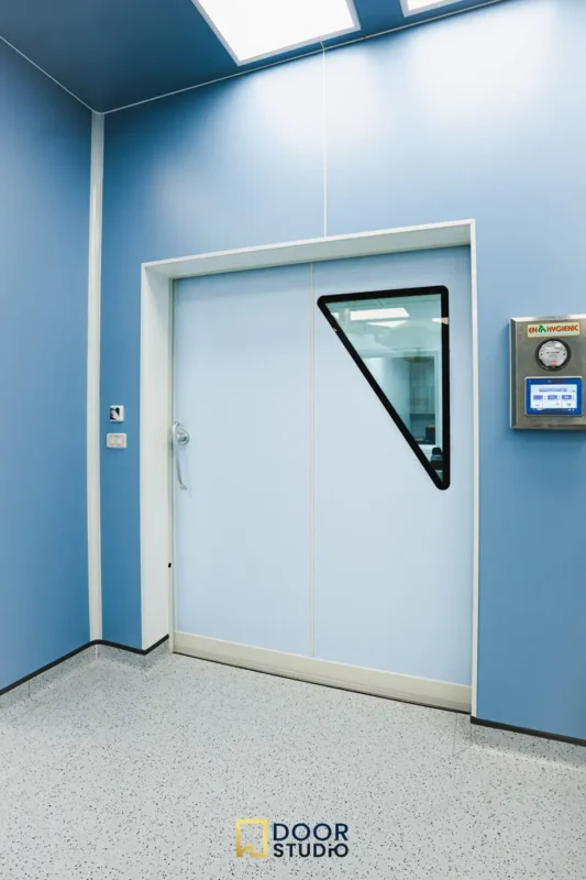 ภาพแสดงประตูห้องผ่าตัดอัตโนมัติแบบบานเลื่อนของ Door Studio ในโรงพยาบาลจุฬาภรณ์ จากมุมมองด้านข้างที่เห็นเพดานห้องด้วย ประตูสีฟ้าอ่อน มีช่องกระจกรูปสามเหลี่ยมที่มุมบน ผนังห้องสีฟ้าอ่อน พื้นเป็นสีเทาอ่อนลายจุด มีอุปกรณ์ควบคุมติดผนัง เพดานมีไฟส่องสว่างแบบฝัง, Image showing a side view with the ceiling of an automatic sliding operating room door by Door Studio in Chulabhorn Hospital. The door is light blue with a triangular glass window at the upper corner. The room walls are light blue, and the floor is light gray with speckled patterns. A control panel is mounted on the wall. The ceiling has recessed lighting.